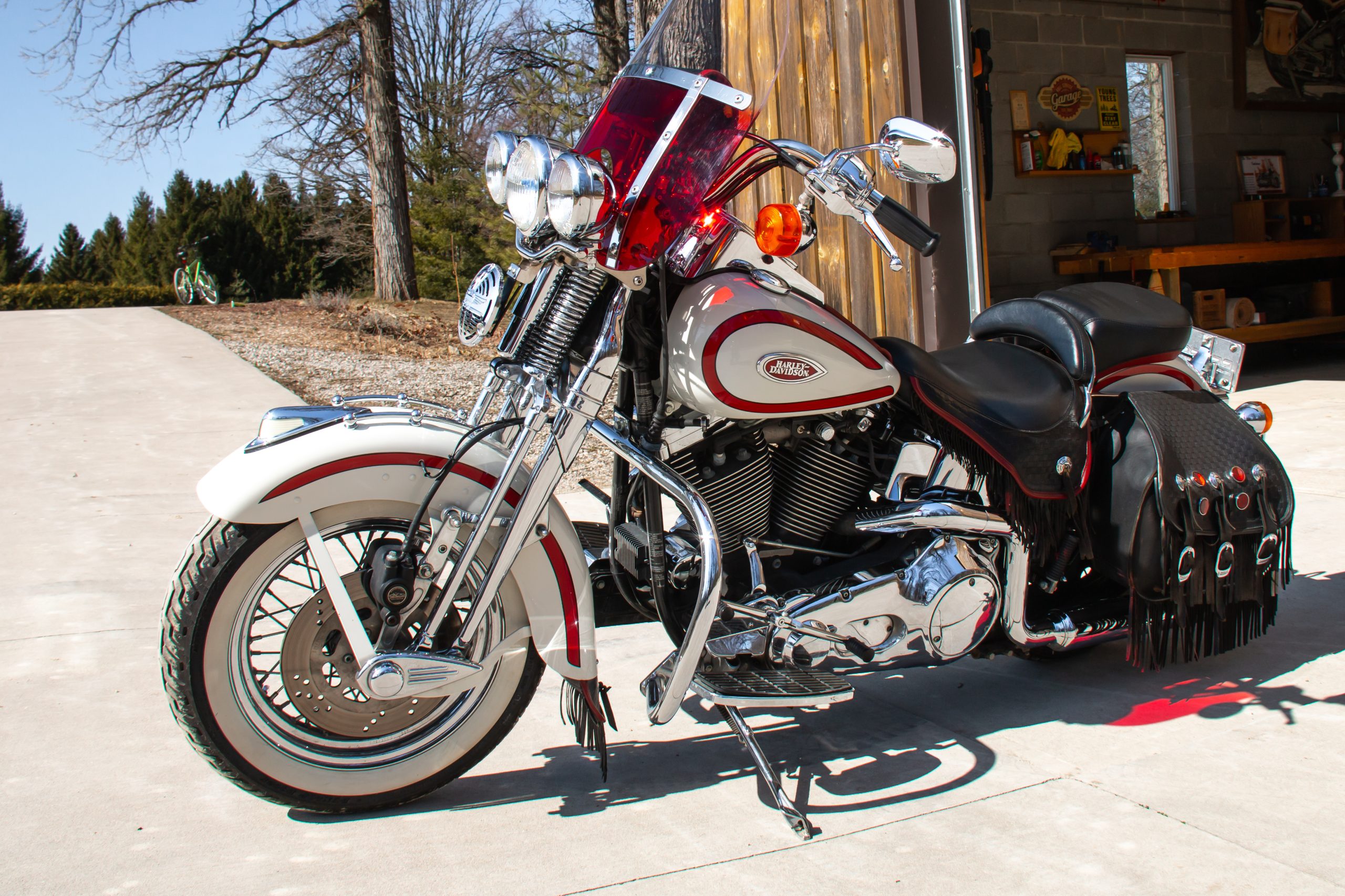 London, Ontario, Canada - March 21 2021: Afternoon shot of a 1996 Harley-Davidson Springer motorcycle in broad daylight with blue skies and spruce trees in the background. Coming out of a brown barn.